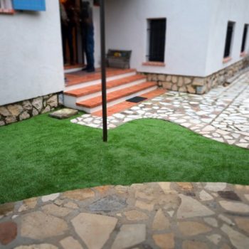 photo_de_entree_de_maison_avec_en_premier_plan_du_carrelage_en_opus_puis_une_partie_en_gazon_synthetique_pôse_par_un_artisan_et_en_dernier_plan_les_marches_en_brique_et_lentree_de_la_maison_et_une_allee_en_opus_devant_des_murs_blancs